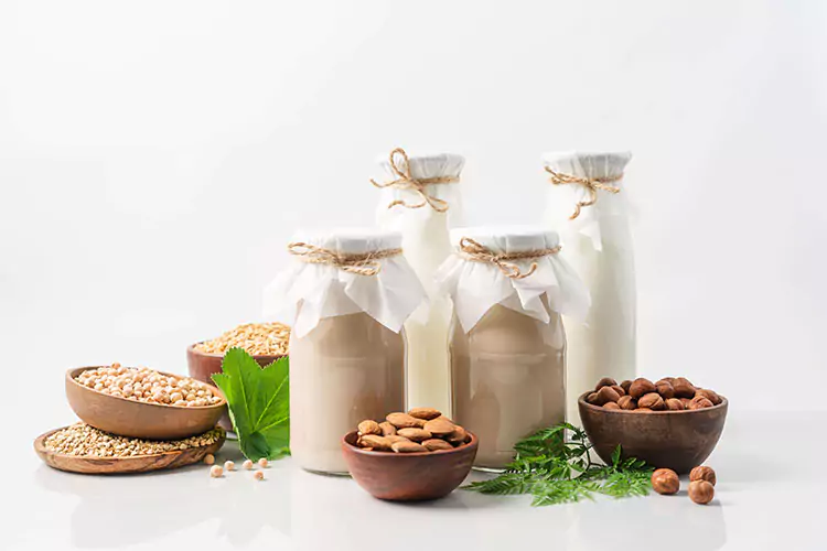 Medium shot of a four glass bottles filled with plant based milk with bowls filled with almonds, macadamia and pecan nuts.